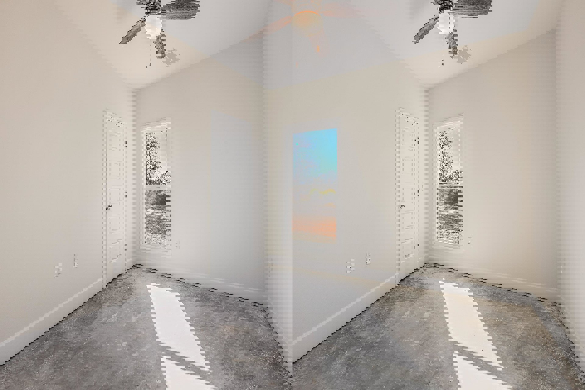 Neutral-toned room with wood flooring, white paneled door with silver knob, ceiling fan with light fixture, and window overlooking leafless trees and a road.