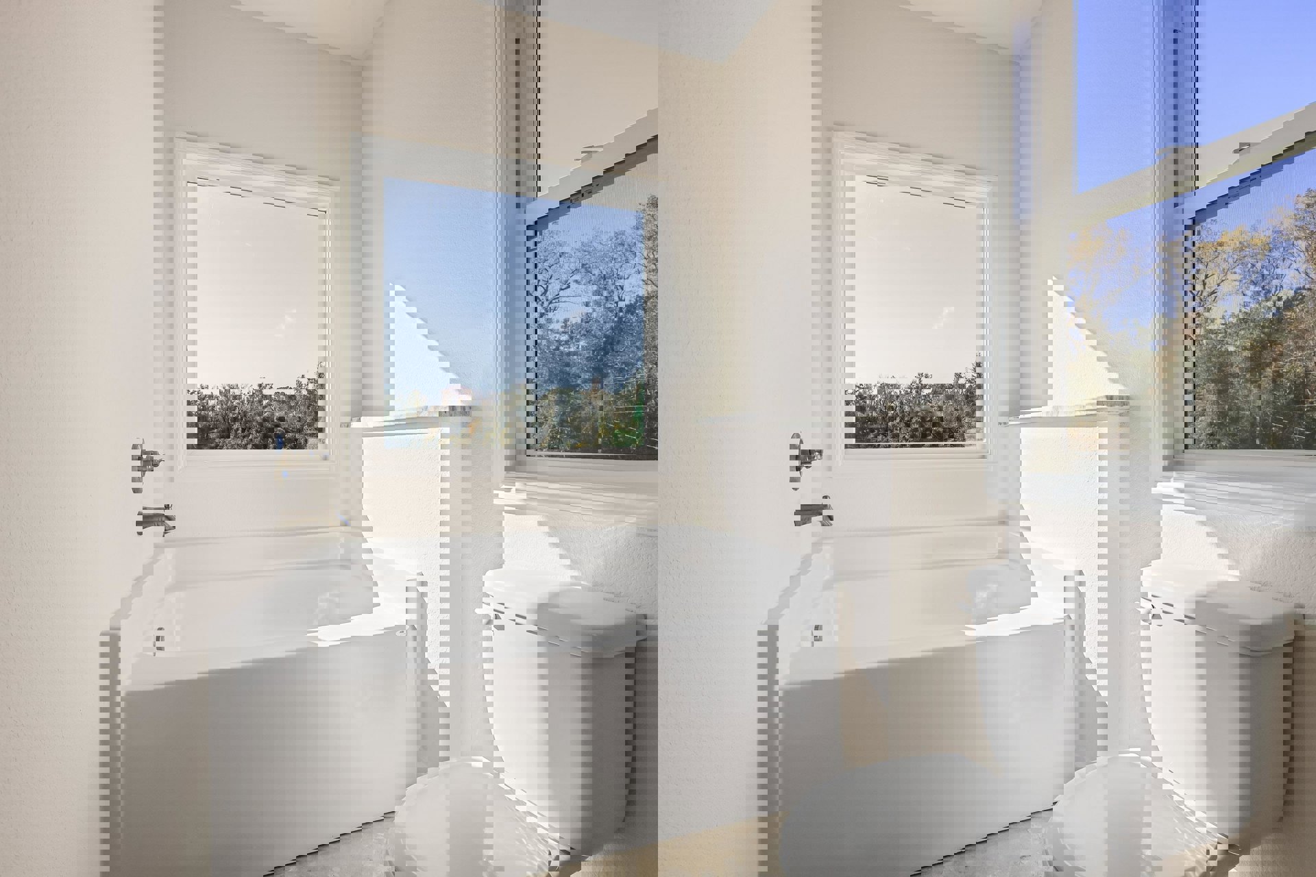 White bathtub with chrome faucet beside a white toilet, large window with screen overlooks green trees, light-colored walls and minimal decor.