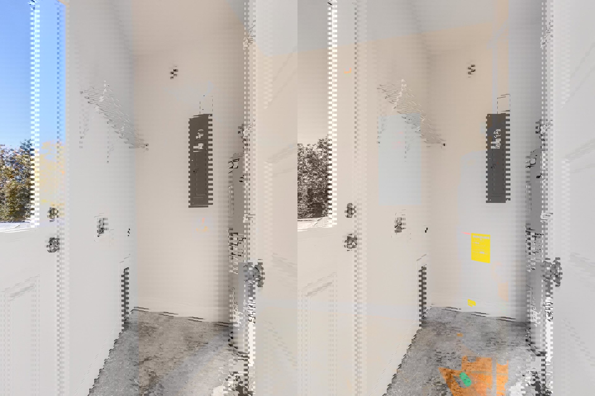 White plaster wall with mounted grey metal cabinet, white metal shelf, grey door with sign, and close-up of silver door knob; sunlight filters through trees outside under blue sky