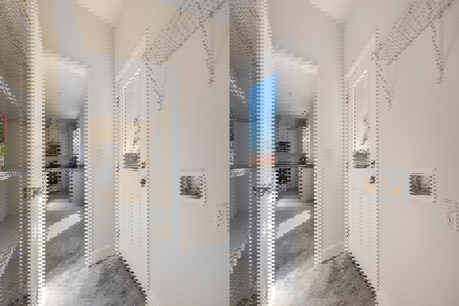 White paneled door beside plaster wall, light wood flooring, built-in cabinetry, stainless oven, electrical outlet, open shelving, window framing tree and blue sky.