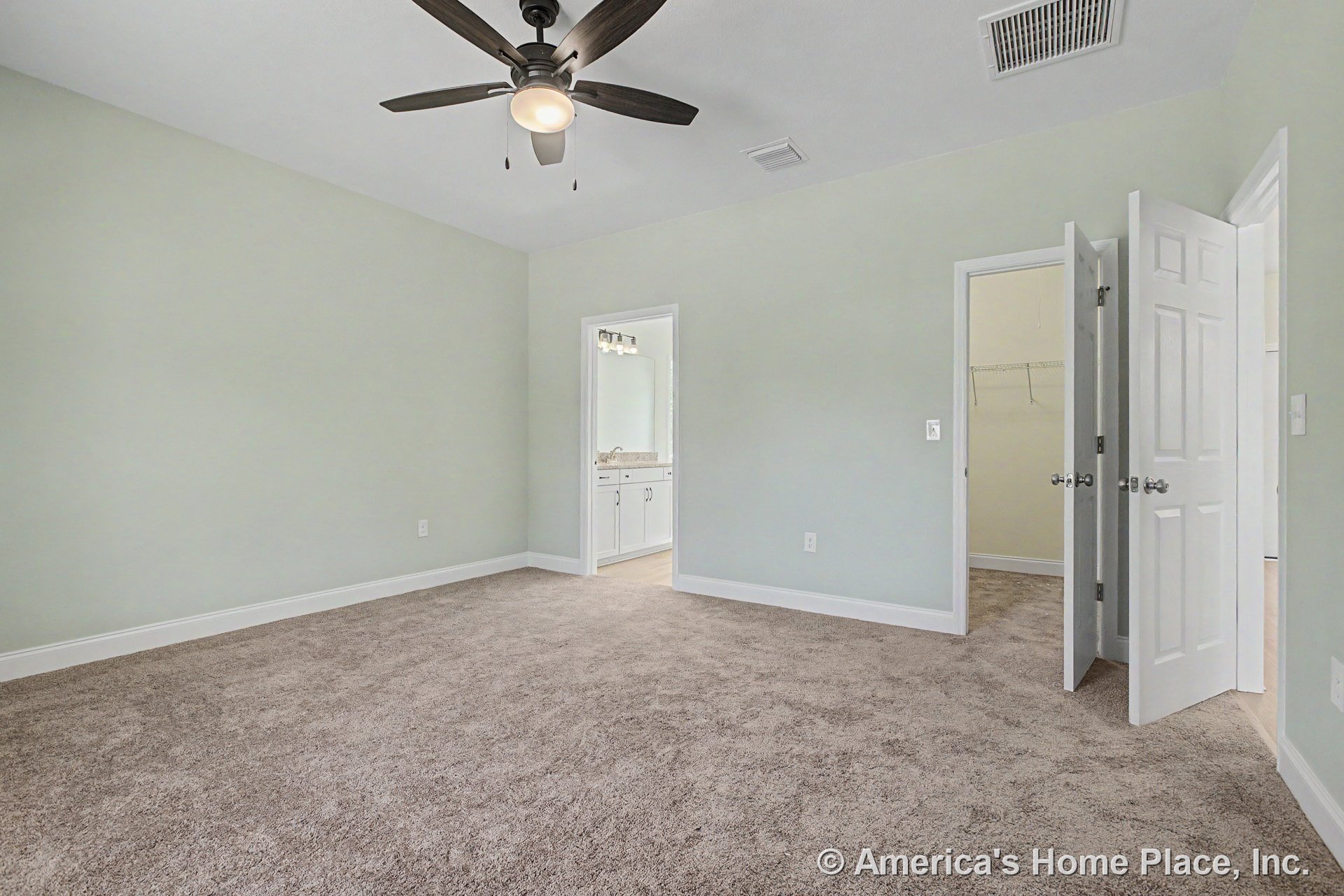 Carpeted bedroom with soft green walls, ceiling fan and built-in light fixture, white baseboards, double closet doors, and direct access to en-suite bathroom featuring white