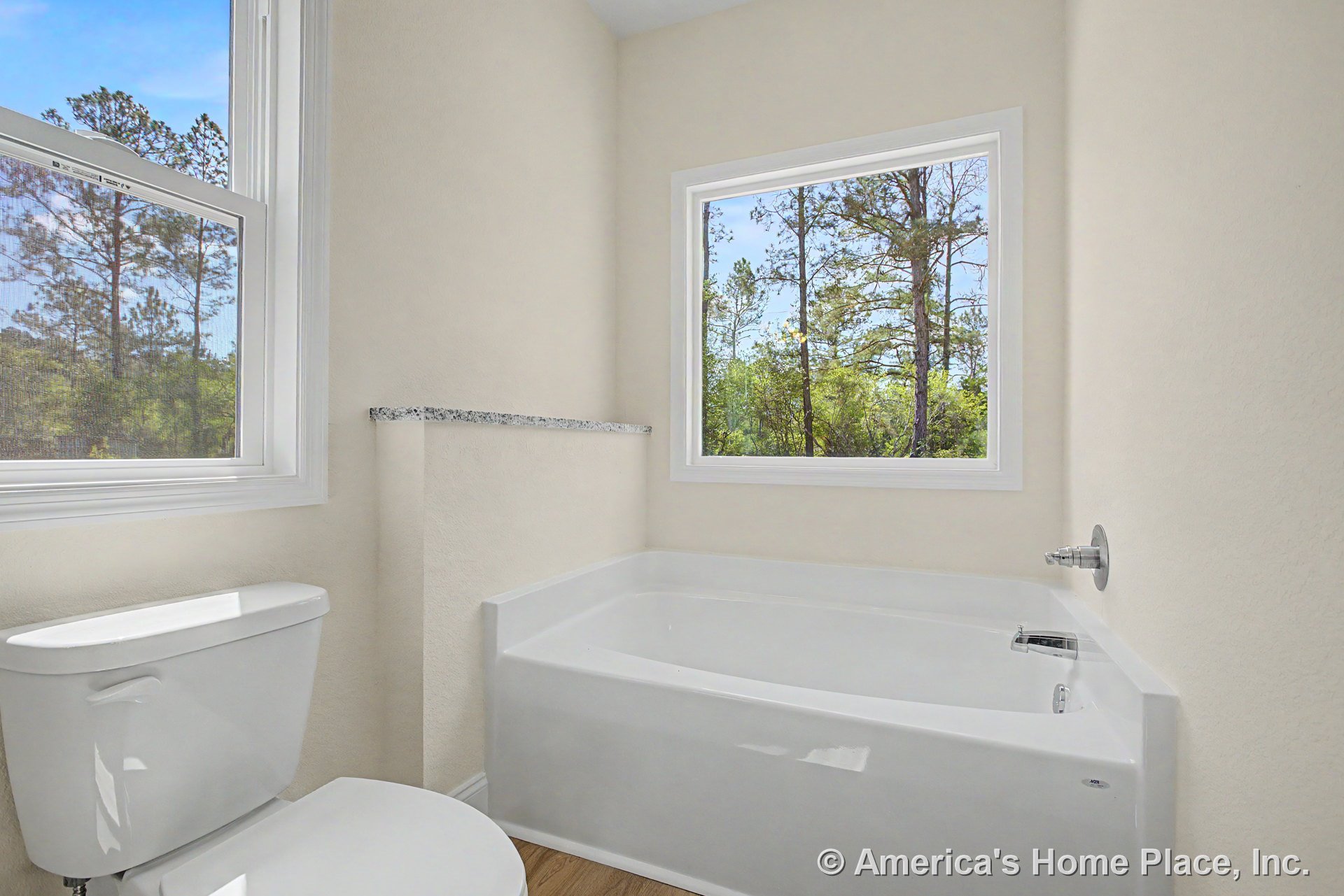 White bathtub and toilet set against neutral walls, large picture window with granite ledge overlooking wooded scenery, wood-look flooring, and abundant natural light.