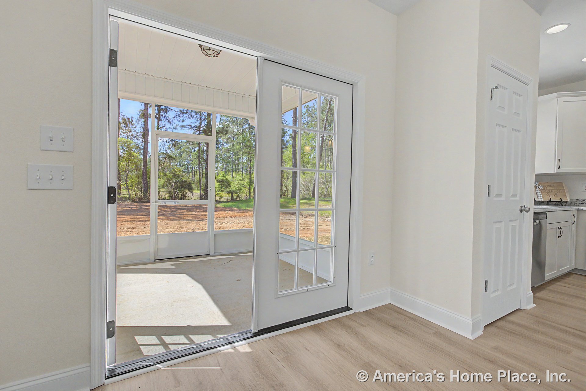 Glass-paneled door leads from kitchen with white cabinetry and light wood flooring onto screened porch featuring ceiling-mounted light, white baseboard trim, and backyard view.