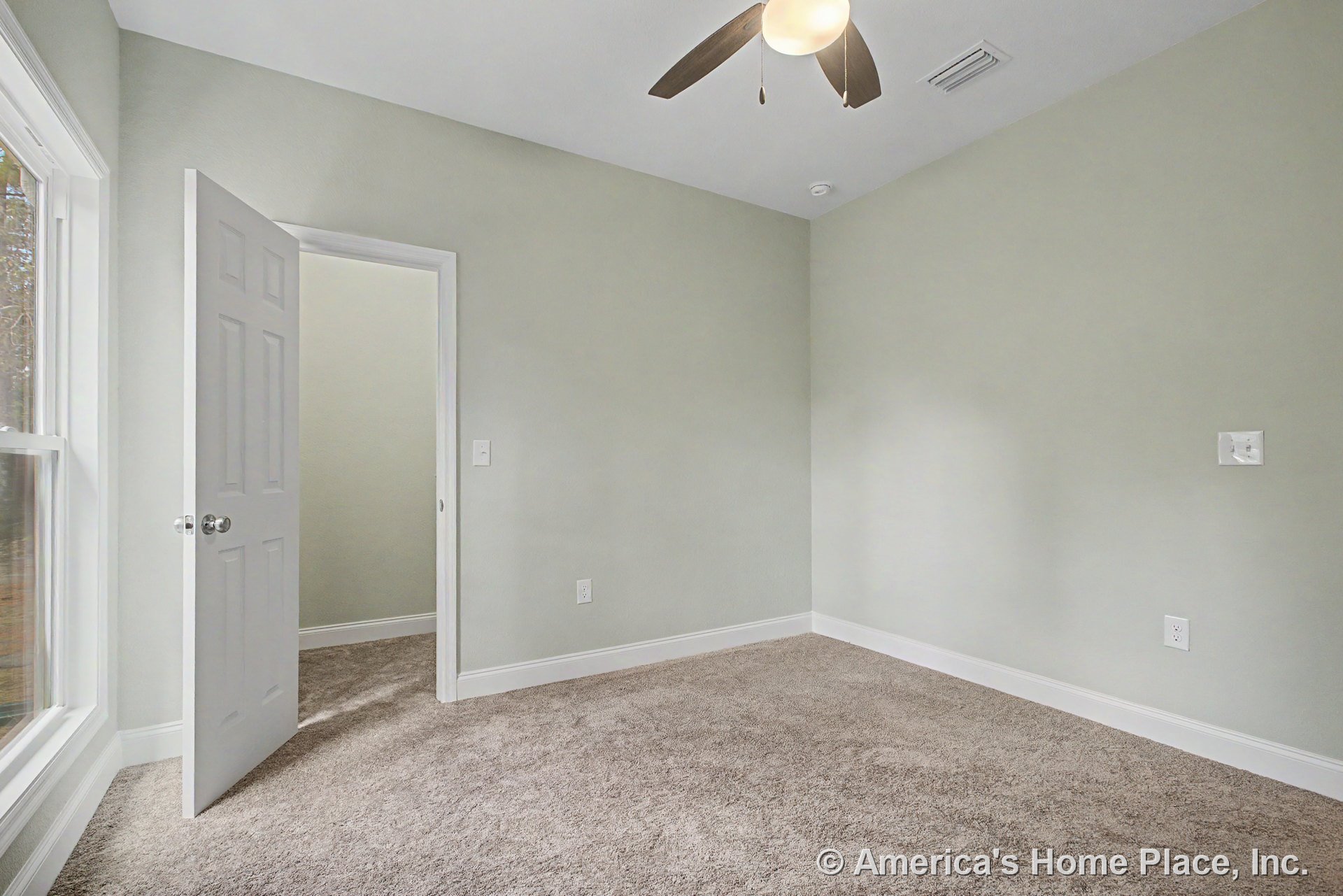 Bedroom with light gray walls, beige carpet flooring, large window with white trim, open white door, ceiling fan with integrated light, and white baseboards.