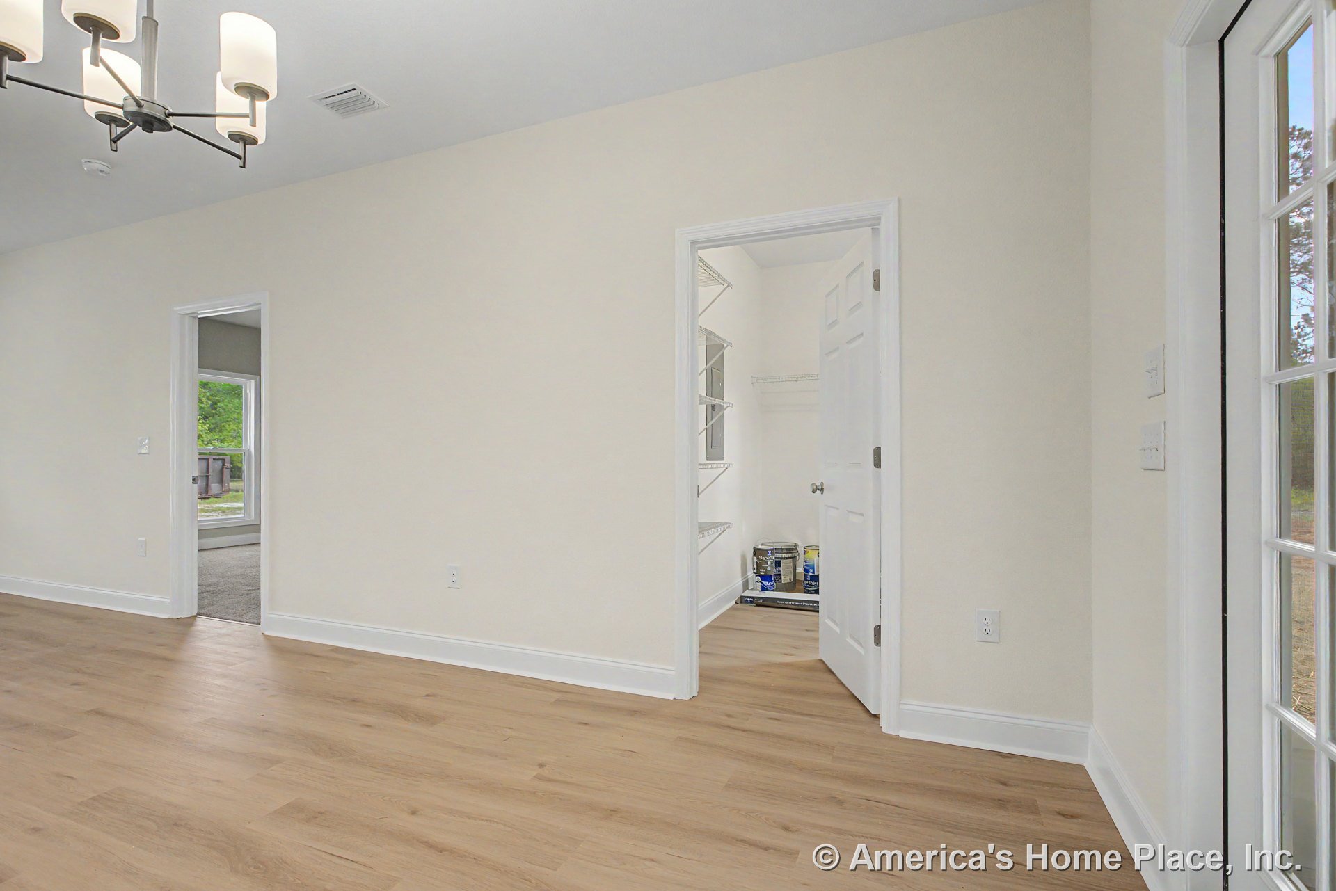 Dining area with light wood flooring, modern chandelier, white trim and baseboards, pantry featuring built-in wire shelving, glass exterior door, and large window in adjacent room.