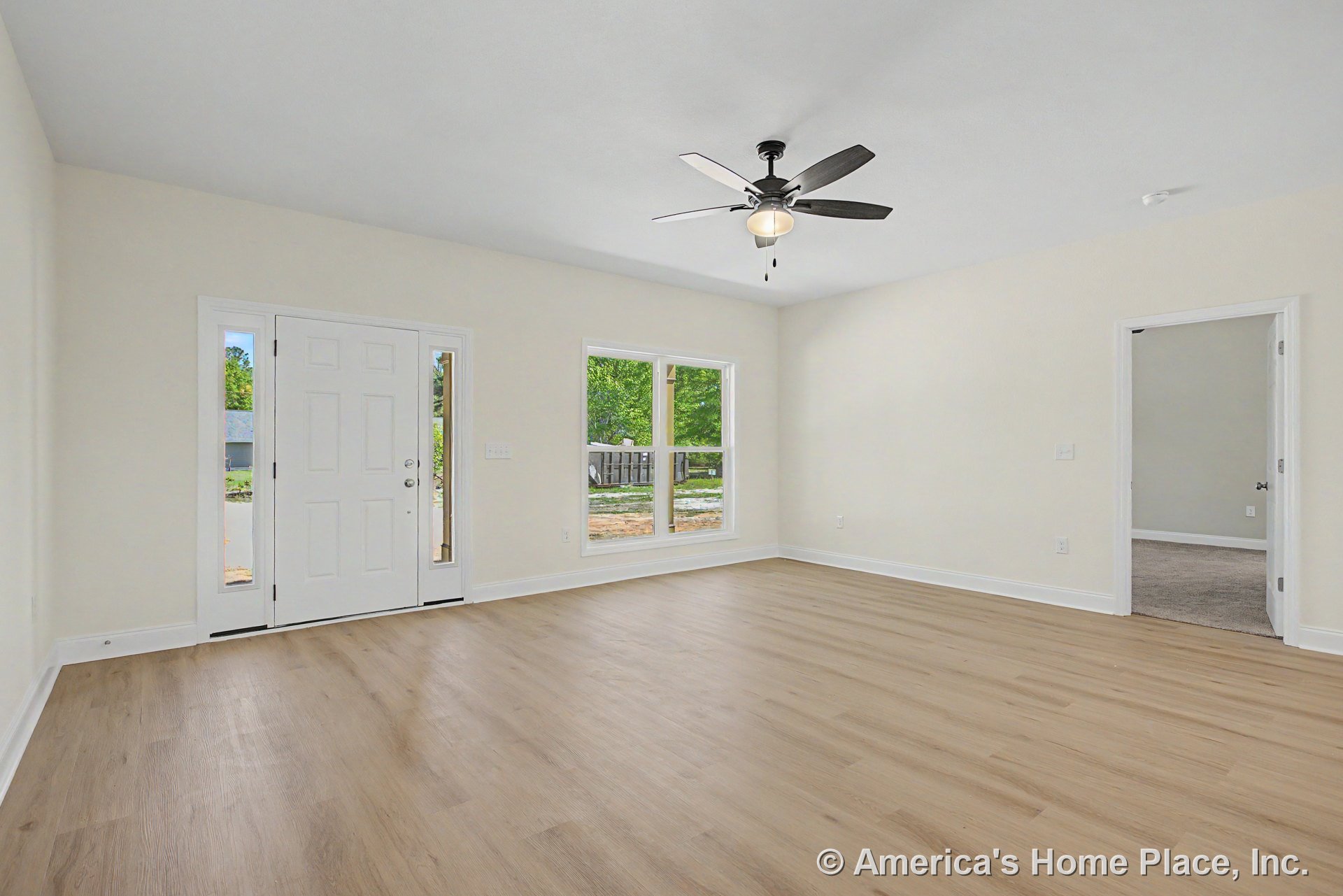 Light wood plank flooring, wide baseboard trim, and neutral wall paint in living room; ceiling fan with integrated light overhead; double window and white front door with