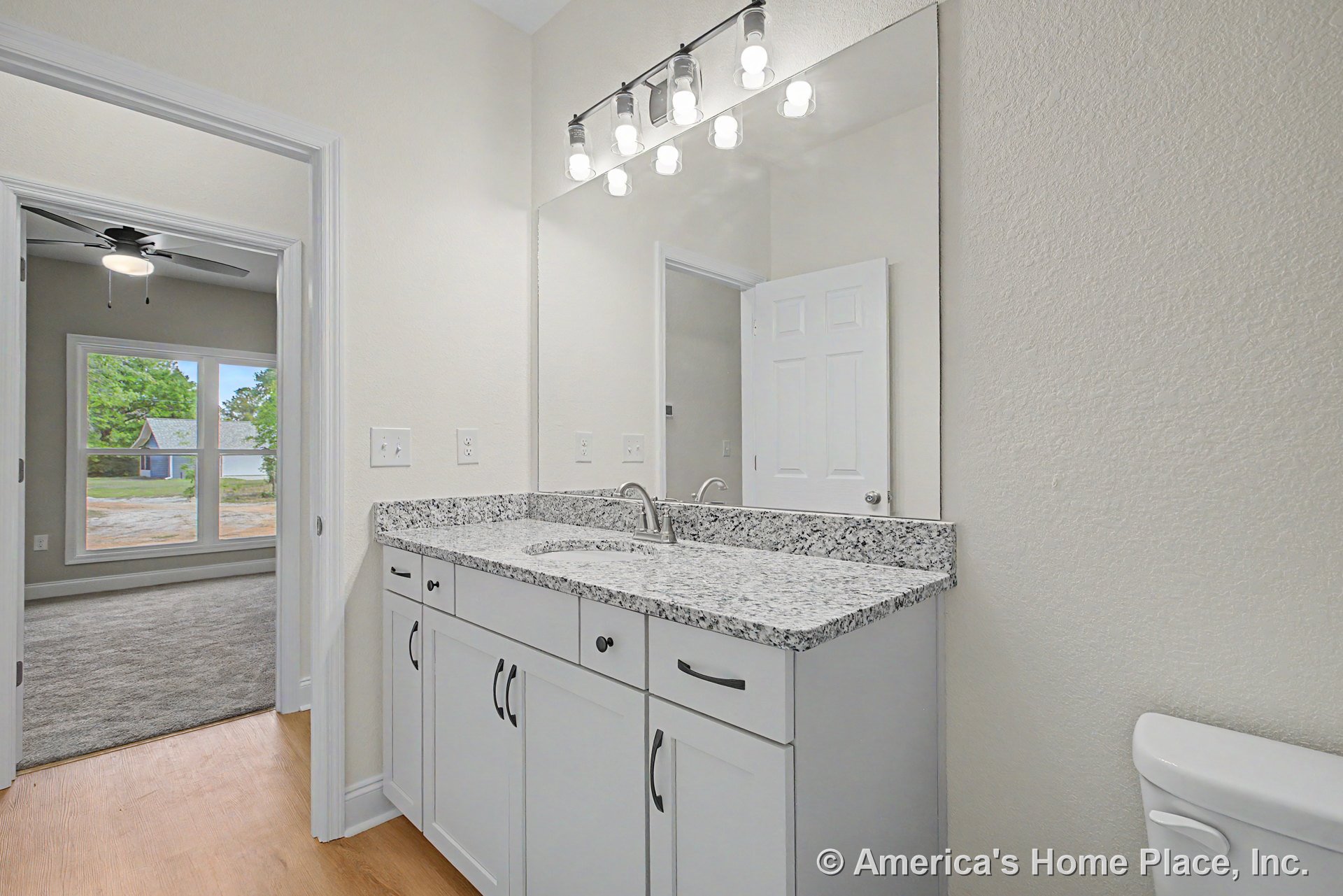 Granite countertop vanity with double sinks, white shaker cabinets, expansive wall-mounted mirror, modern light fixture, wood-look flooring, white trim, and window adjacent to