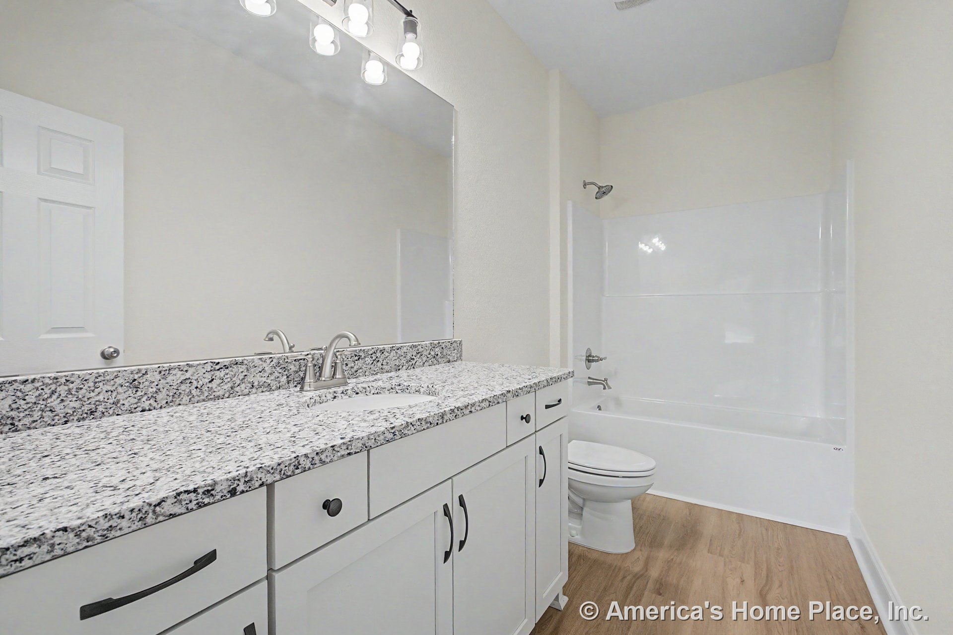 Granite vanity with single chrome faucet, white shaker cabinets, expansive wall mirror, built-in overhead lighting, integrated tub-shower combo, wood-look flooring, and neutral