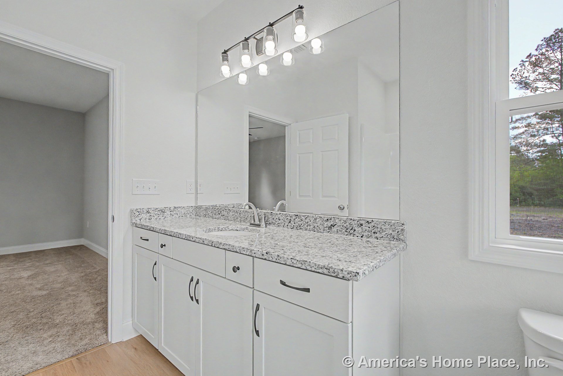 Granite countertop with single undermount sink, white shaker cabinets, expansive wall mirror, wall-mounted vanity light fixture, and window with decorative trim.