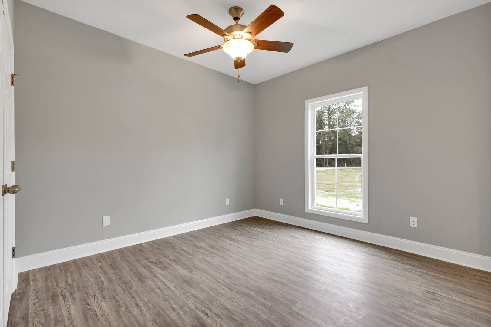 Ceiling fan with light fixture mounted on white plaster ceiling, large window overlooking green lawn and tree with power lines, wood laminate flooring with white baseboards