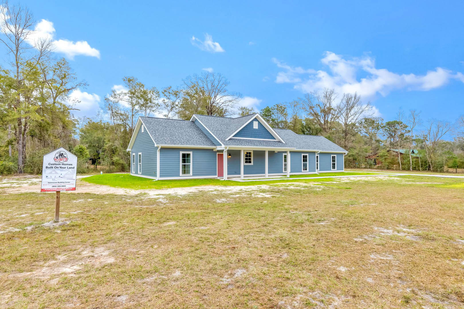 Blue siding house with red front door, expansive green lawn, wooden post sign near driveway, cluster of mature trees and blue sky in background