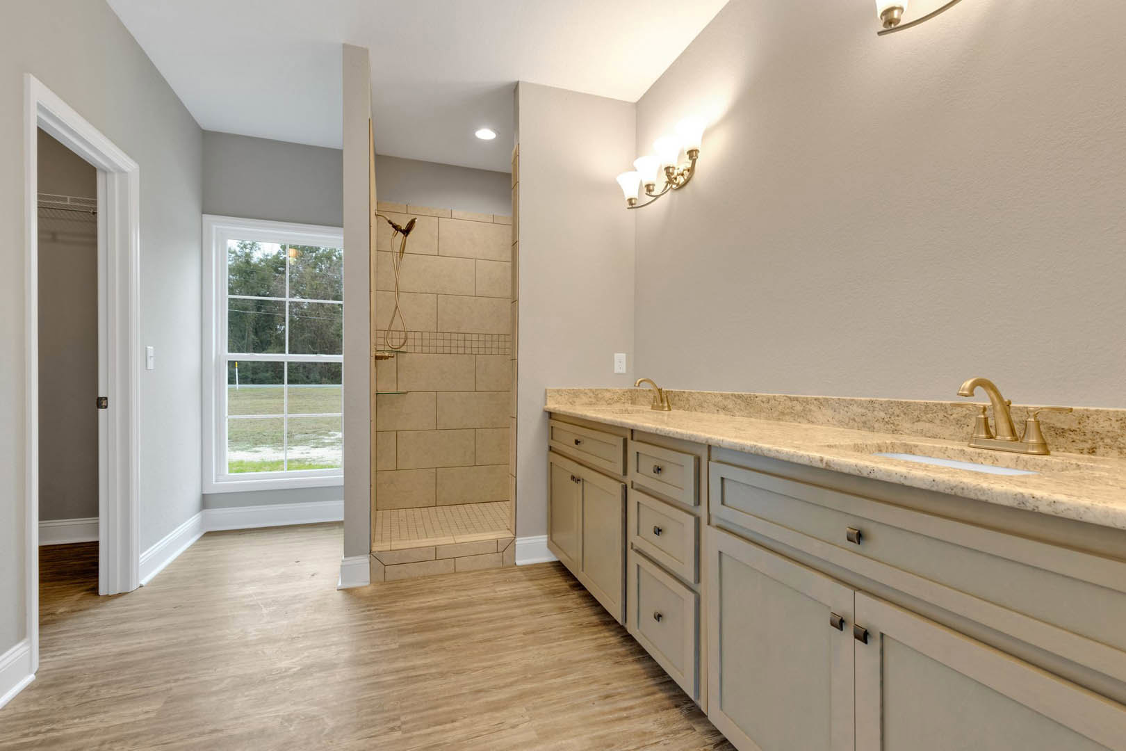 Bathroom featuring a glass-enclosed tile shower, white sink with stone countertop, wood cabinetry, and a window overlooking a grassy field; transition from wood to tile flooring
