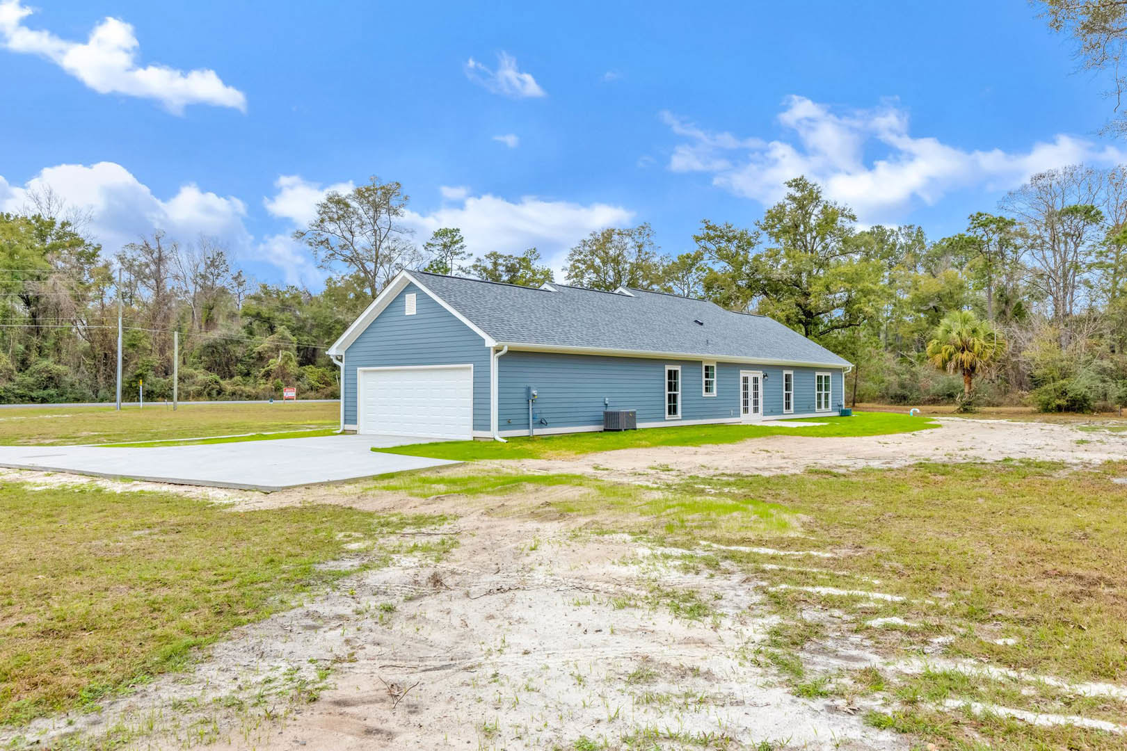 Blue house with white garage door, blue siding, and driveway; dirt yard in front, palm tree and wooded area in background, partly cloudy sky overhead.