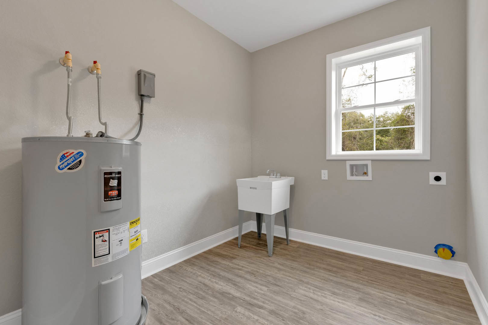 Utility room featuring a white water heater with stickers, square sink with silver faucet, wood flooring, table and chair, and window overlooking trees.