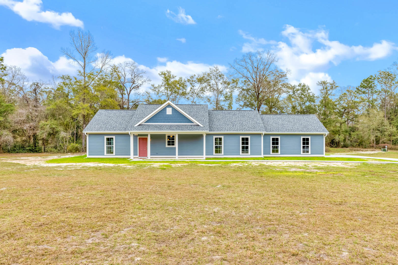Blue-roofed farmhouse with white vent, surrounded by a large grassy lawn, wooden fence, mature trees, and under a partly cloudy sky