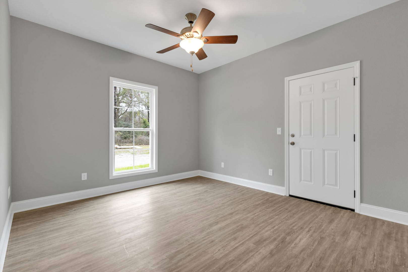 Ceiling fan with light fixture mounted on white ceiling, large window overlooking trees and grass, wood flooring with white baseboard trim, white door with silver handle