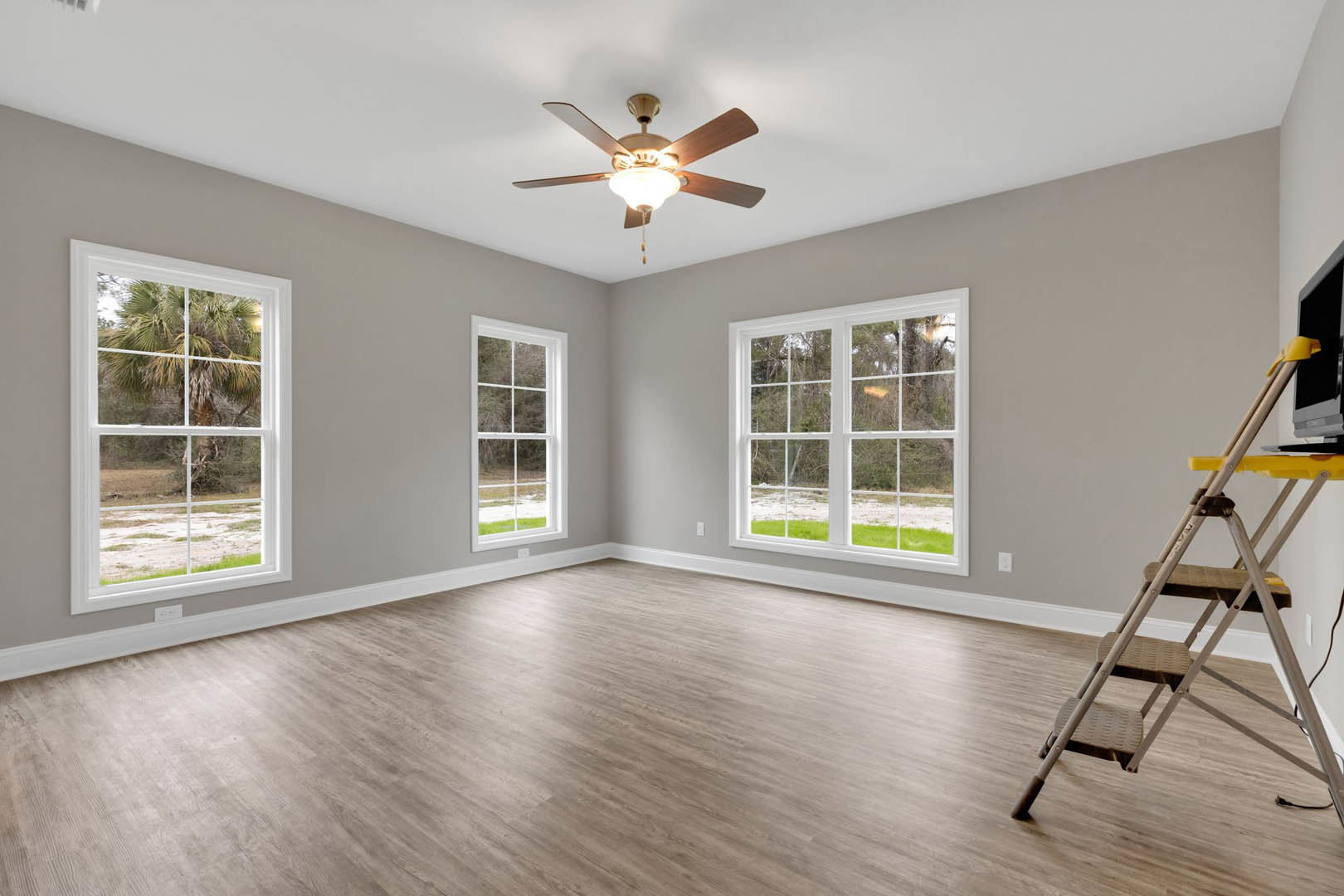 Living room with wood laminate flooring, white plaster walls, ceiling fan with light, metal ladder beside wall-mounted television, large window framed in white showing palm tree