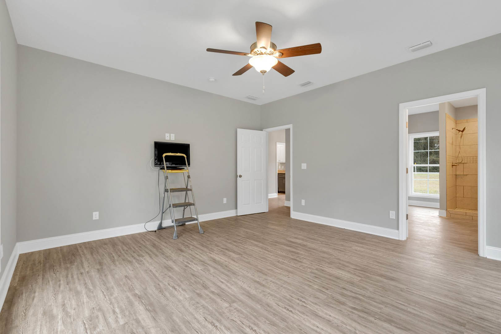 Hardwood floor room with white walls, ceiling fan with light fixture, white door, and metal ladder topped with a black bag