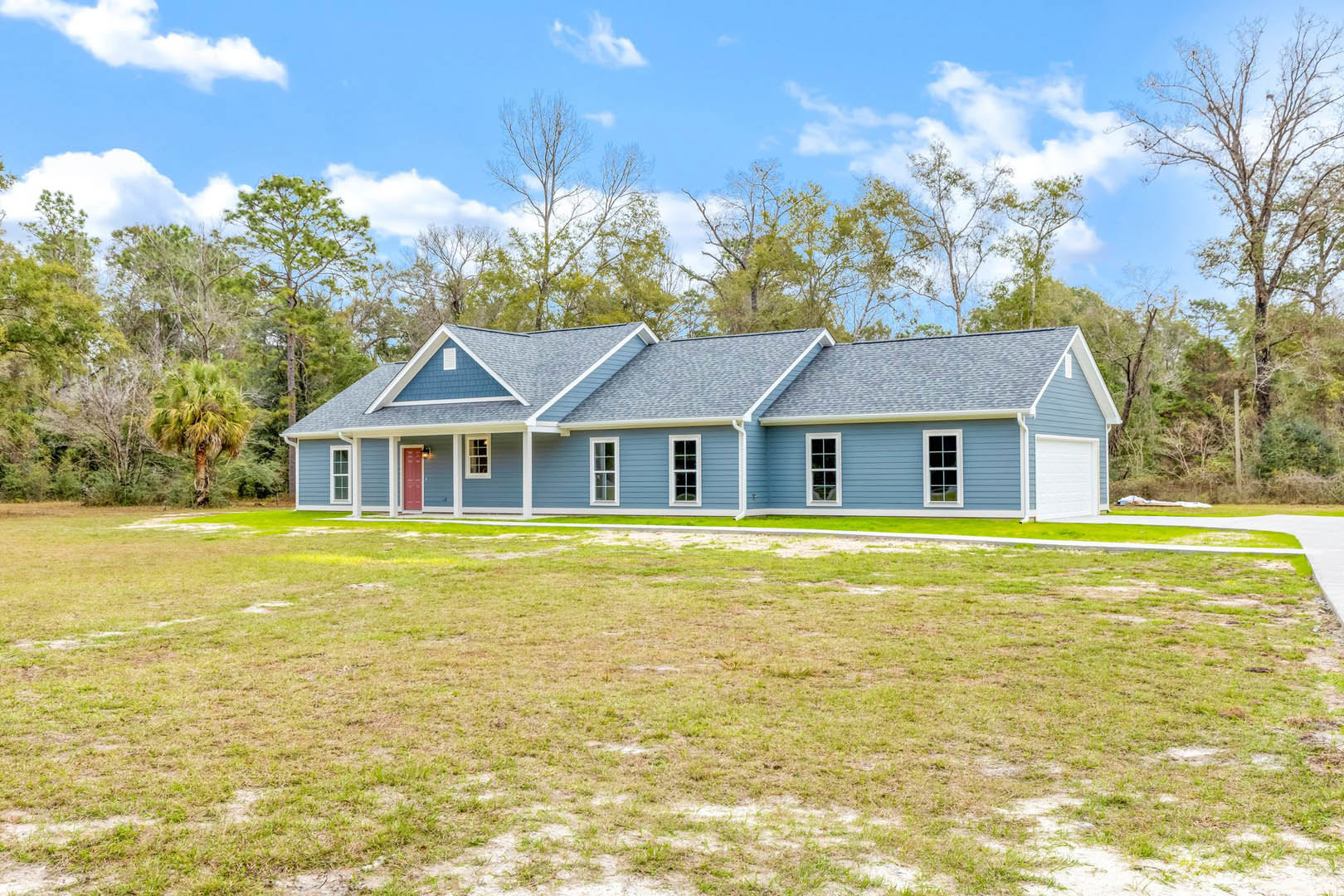 White house with attached garage, surrounded by green grass lawn, palm tree and other mature trees under blue sky with scattered clouds