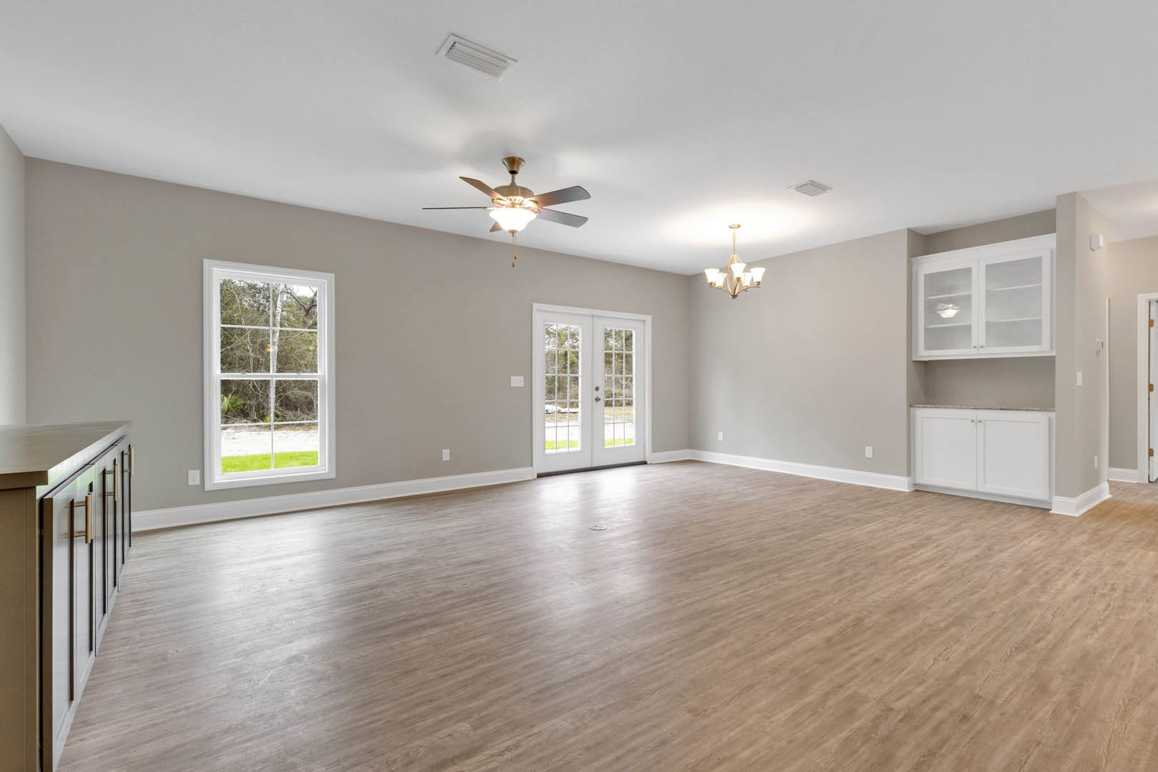 Spacious room with hardwood flooring, ceiling fan with light fixture, double glass-paneled doors, large window overlooking trees, and white cabinet with glass doors