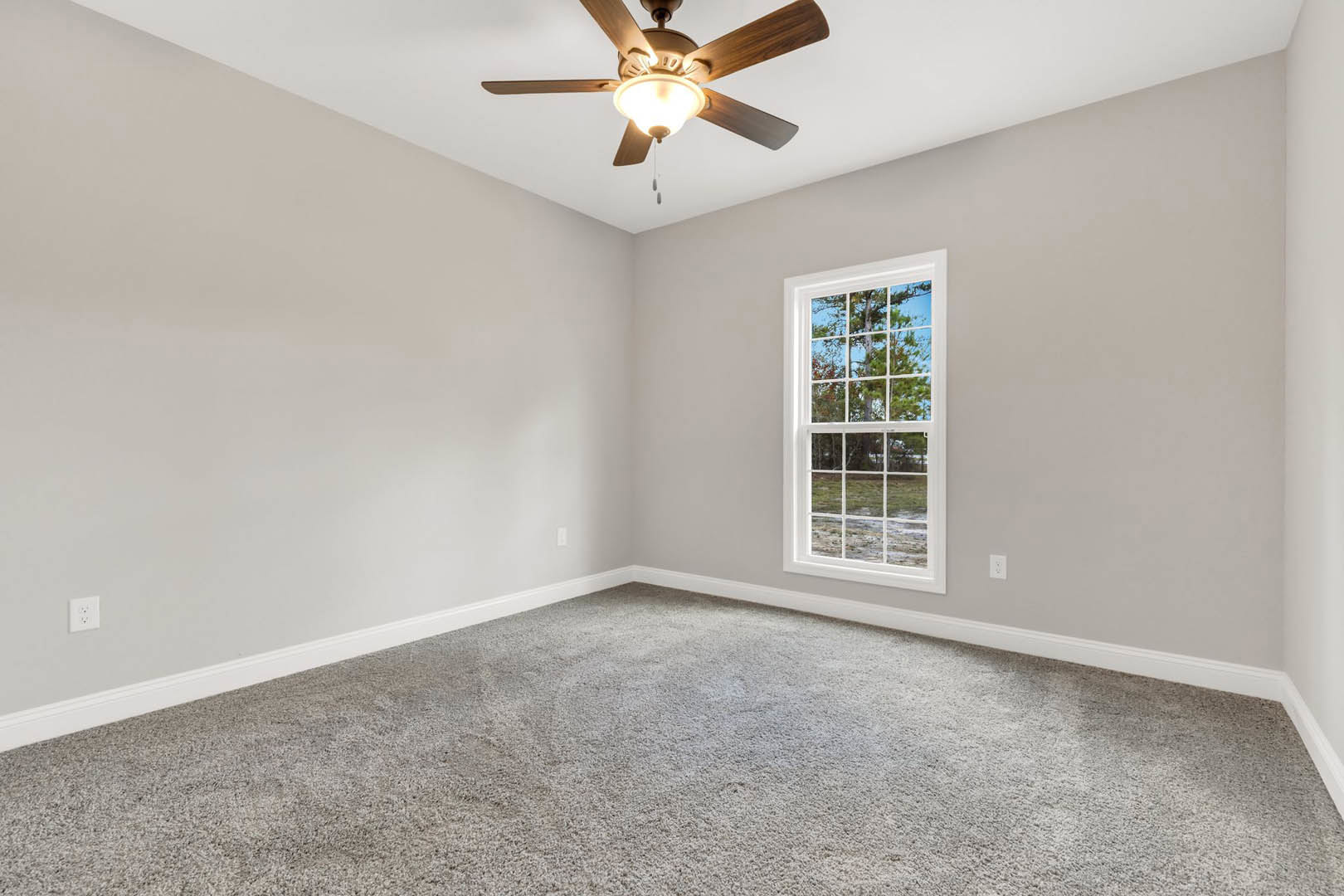 Neutral-toned carpeted room with white walls, ceiling fan and light fixture, large window framed by molding, view of green trees outside