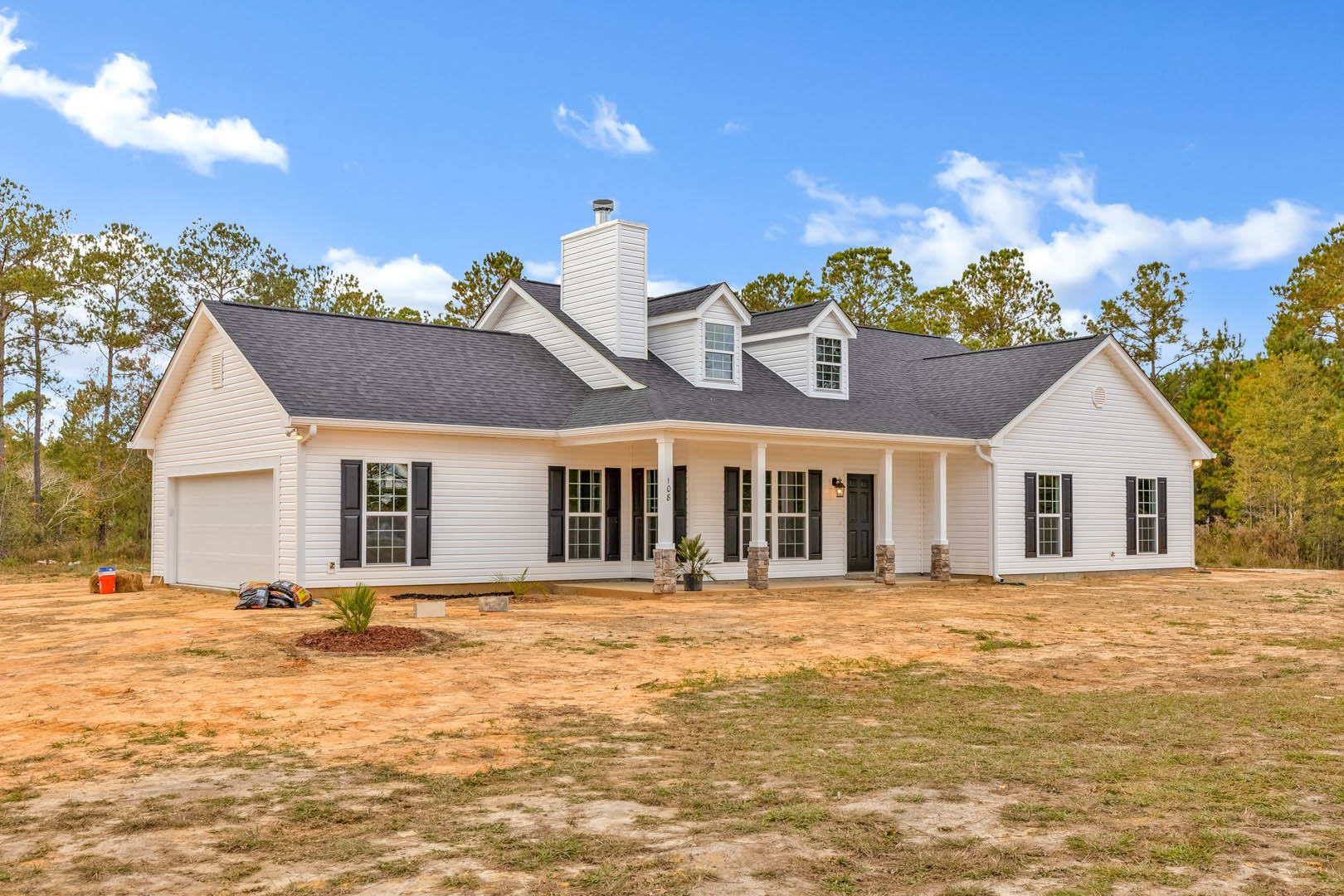 White siding house with black shuttered windows, brick chimney, expansive dirt yard, and mature trees in the background