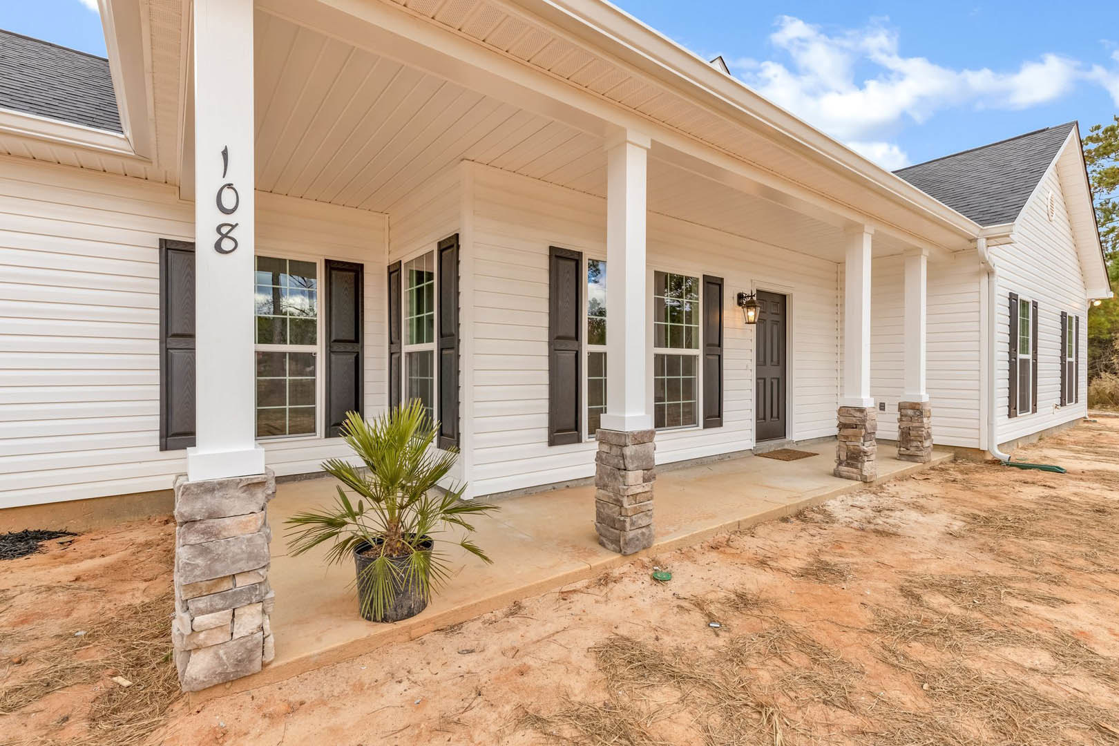 Front porch with stone columns, stacked brick pillars, potted plants, and palm tree beside light-colored siding and windows.