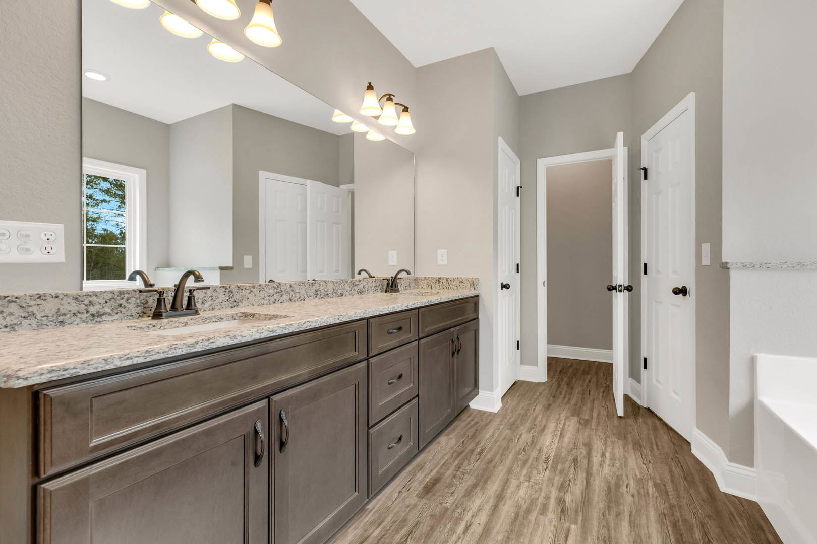 Bathroom with marble countertop, wood floor, white cabinetry, black faucet, white door with black handle, window showing trees outside, white trim along baseboards