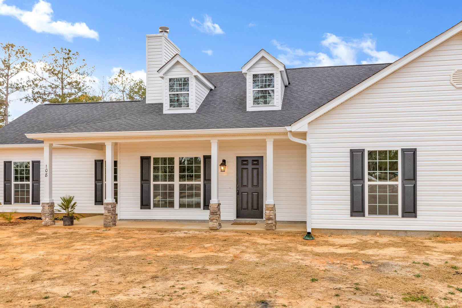 White siding house with grey roof, black front door, black shuttered windows, potted plant on porch, dirt and grass landscaping