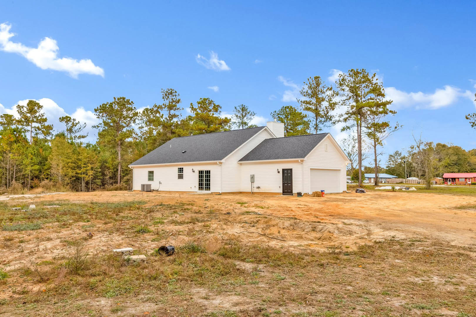 White cottage-style house with multi-pane windows, gray shingle roof, and dirt field in foreground; trees and cloudy sky in background, lawn mower near side of house.