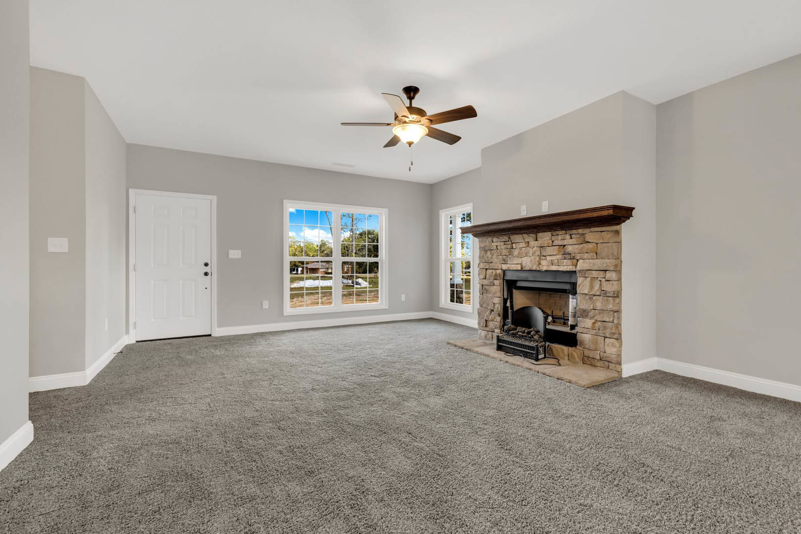 Living room with beige carpet, brick fireplace topped by a wood mantel, ceiling fan with light fixture, white door with black hardware, window overlooking green yard.