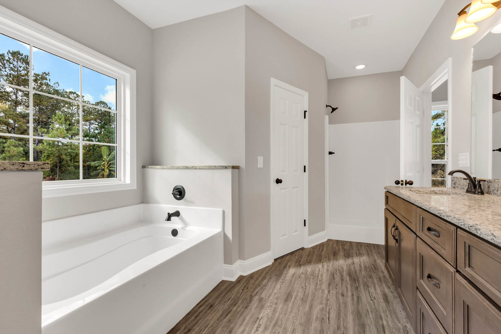 White freestanding bathtub with matte black faucets beside a modern sink, white cabinetry, and light fixture; large window frames view of green trees, white door with silver