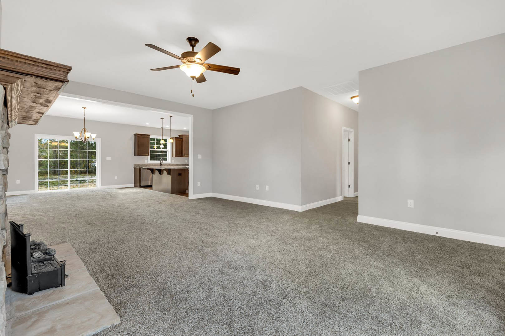 Spacious living room with carpet flooring, ceiling fan with light, open kitchen featuring black stove and brown wooden cabinets, large window offering outdoor view, white walls and
