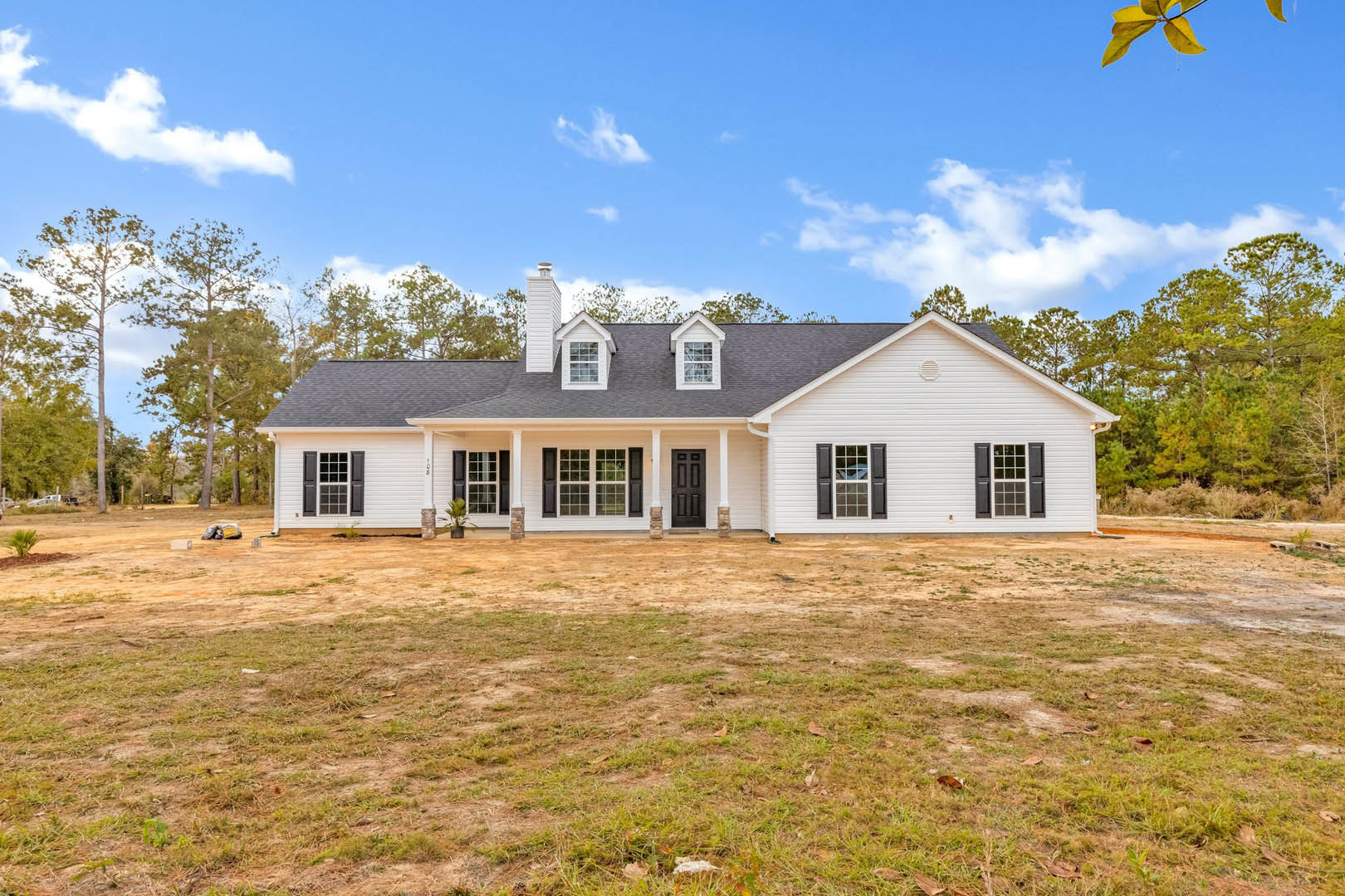White two-story house with black front door, white-framed windows, prominent chimney, surrounded by large grassy yard and mature trees under partly cloudy sky