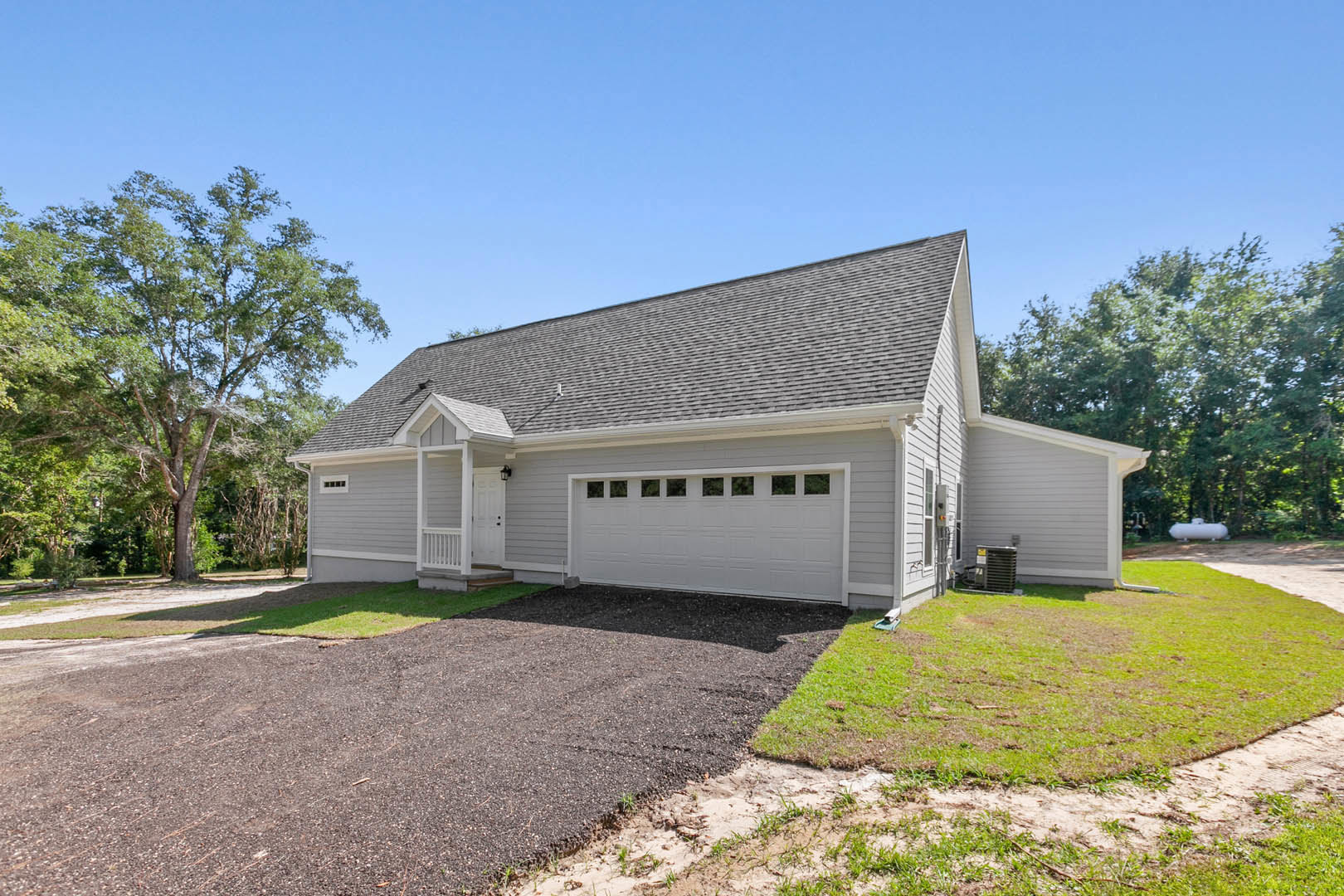 Two-story house with white garage door and windows, covered front porch with white railing, paved driveway, mature trees, manicured lawn, and blue sky with scattered clouds
