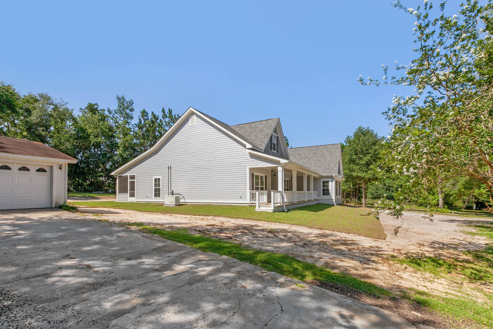 White house with brown roof, attached garage, white porch, concrete driveway bordered by green grass, mature trees including one with white flowers, blue sky overhead
