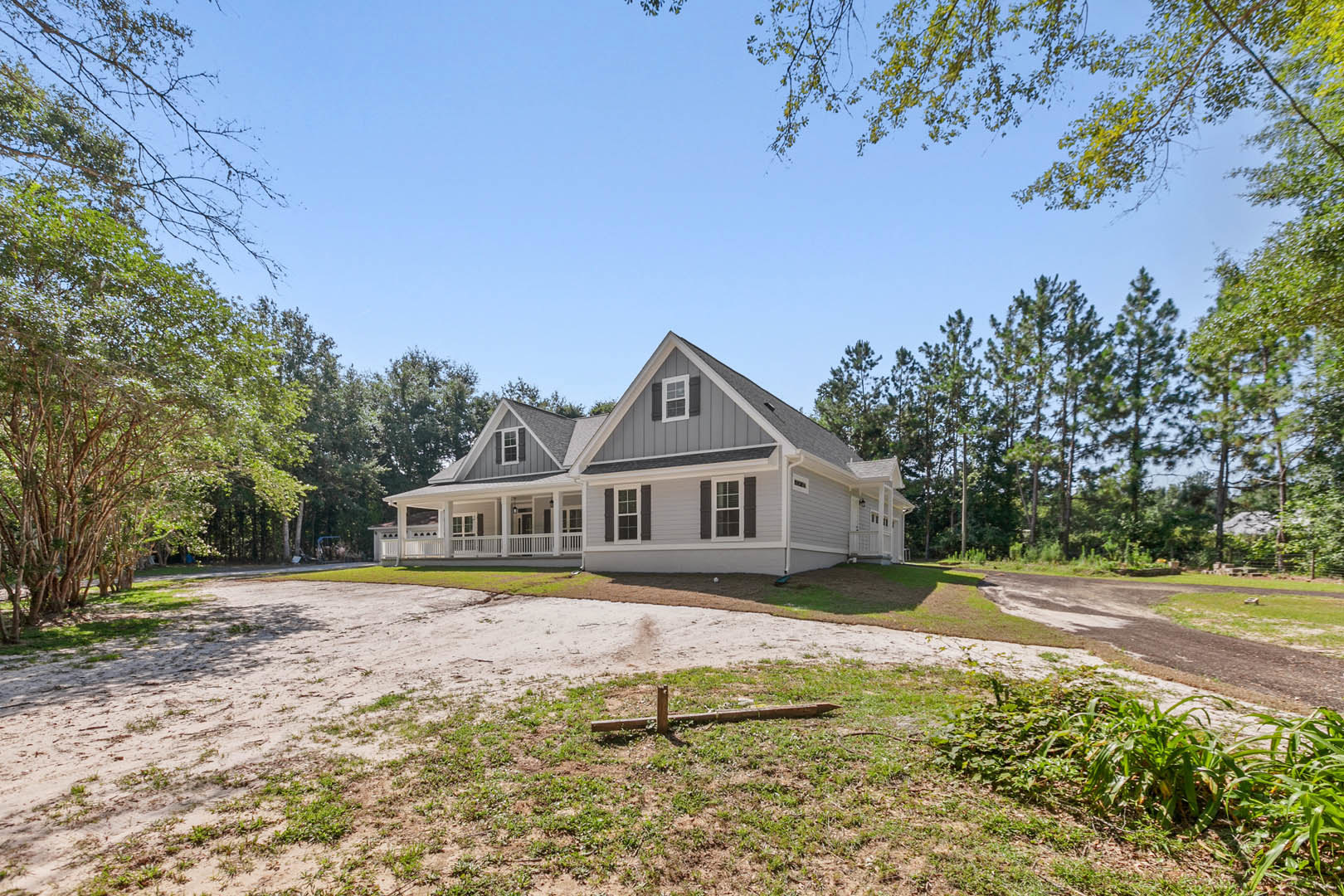 Two-story custom home with stone and siding exterior, spacious concrete driveway, covered front porch, manicured lawn, wooden fence post, mature trees, and landscaped plant beds