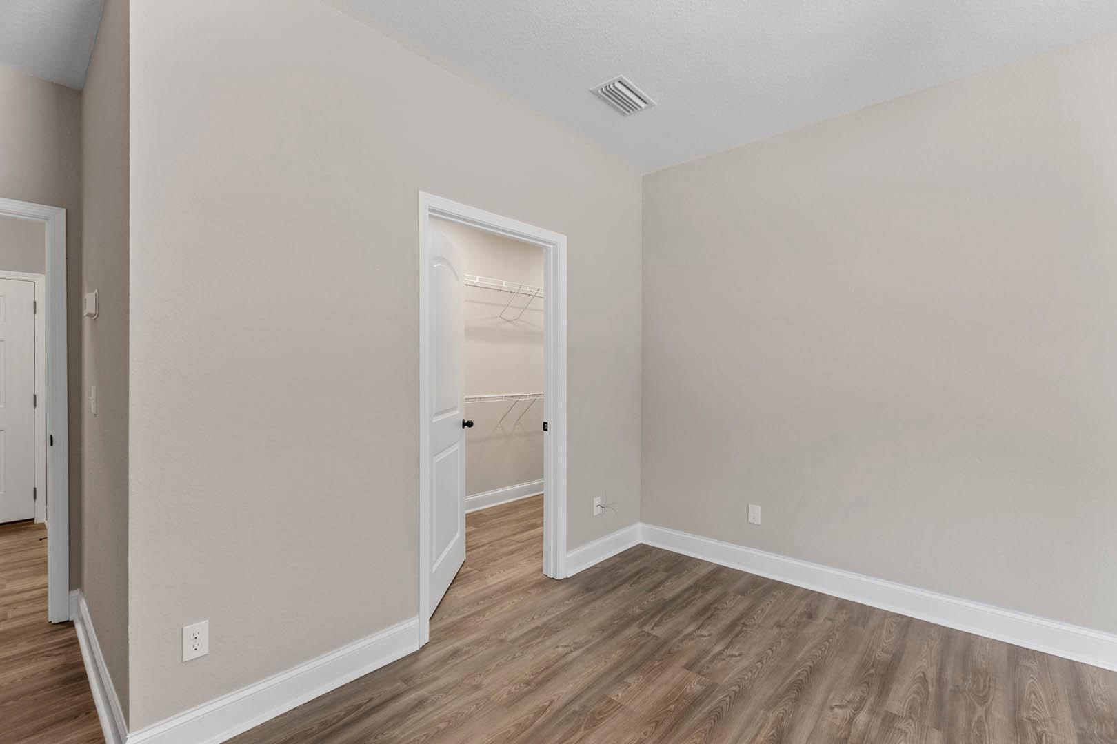 Open white door with black handle, wood flooring with white baseboard trim, white walls, ceiling vent visible