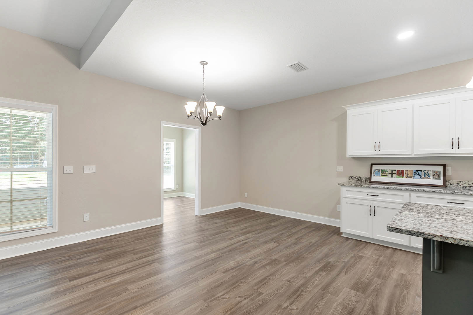 Wood flooring, white cabinetry, window with blinds, picture frame collage, chandelier hanging from white ceiling, kitchen countertop