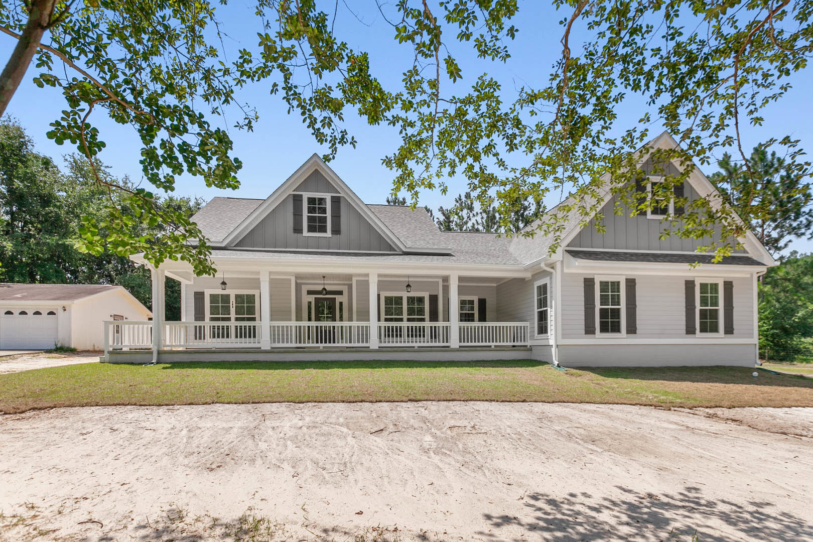 Spacious front porch with white railings, large windows with white frames, sandy landscaping, tree branches overhead, white garage and fence