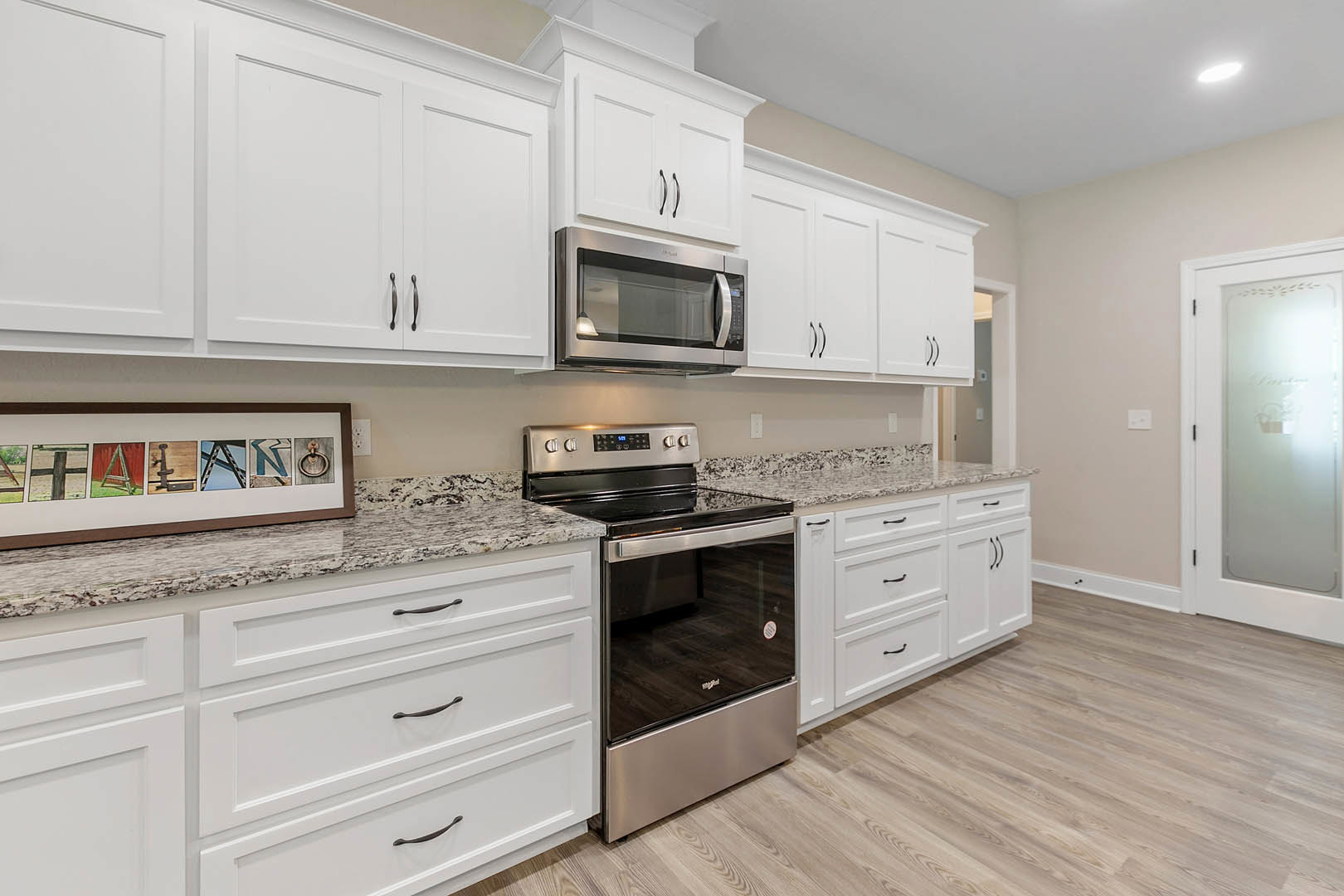 White kitchen cabinets with stainless steel stove, built-in microwave, light countertops, and frosted glass door displaying framed pictures on adjacent wall