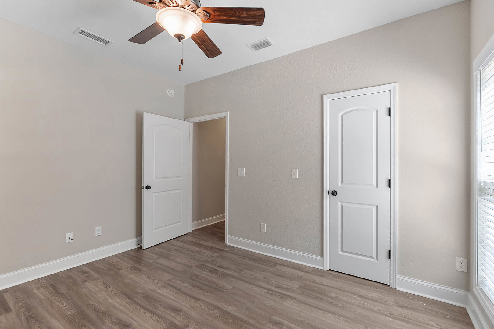 Wood floor with white trim, two white doors with black knobs, ceiling fan with light, white vent on white ceiling