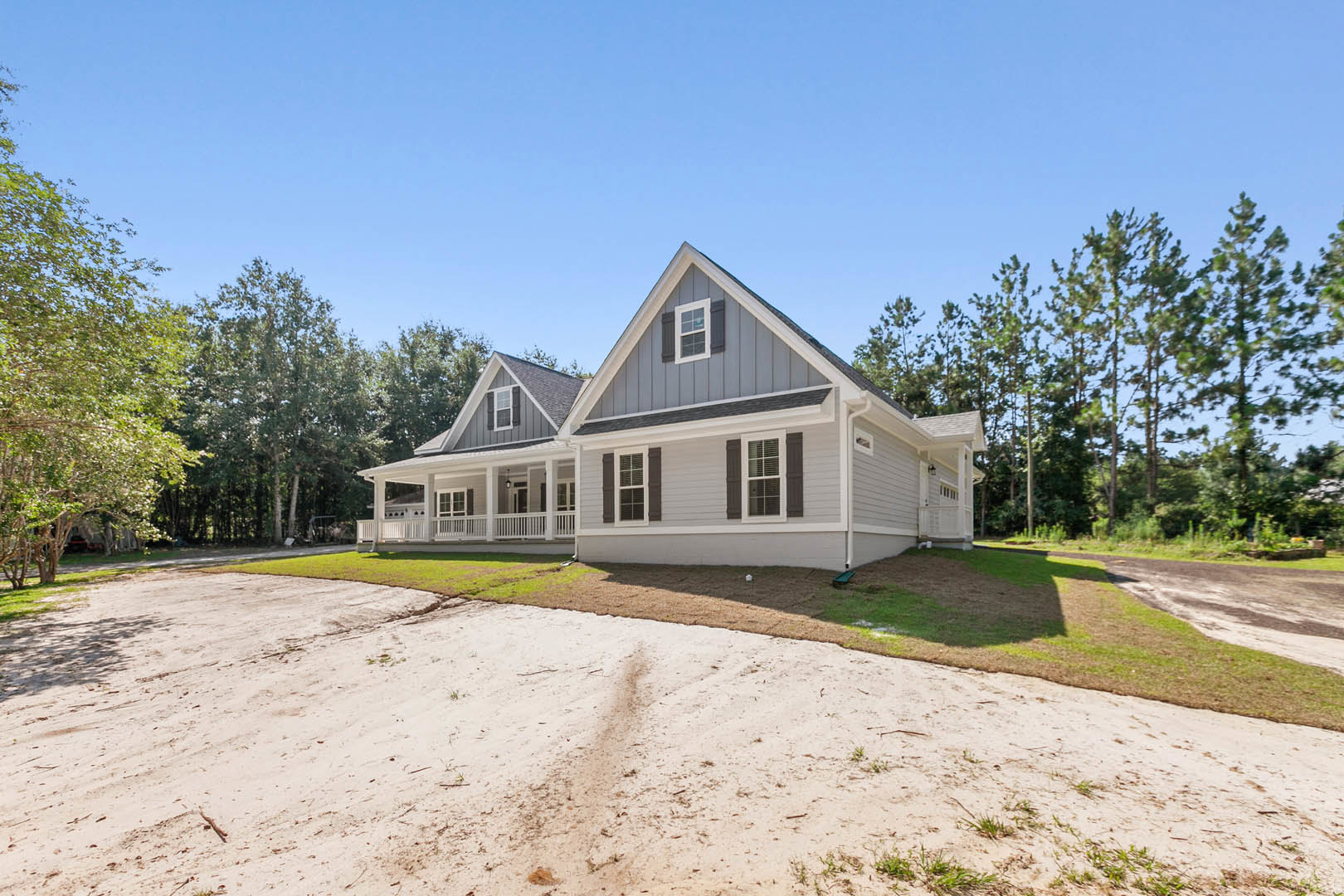 Two-story home with covered front porch, concrete driveway, and mature trees in the background; dirt patch and lawn in front yard.