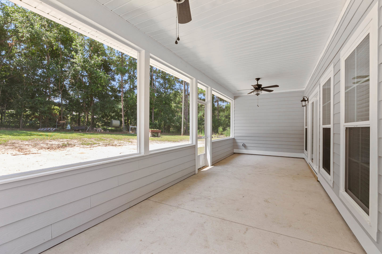 Spacious white interior room featuring multiple windows, white flooring, ceiling fan with light fixture, and a view of porch with trees through glass doors.