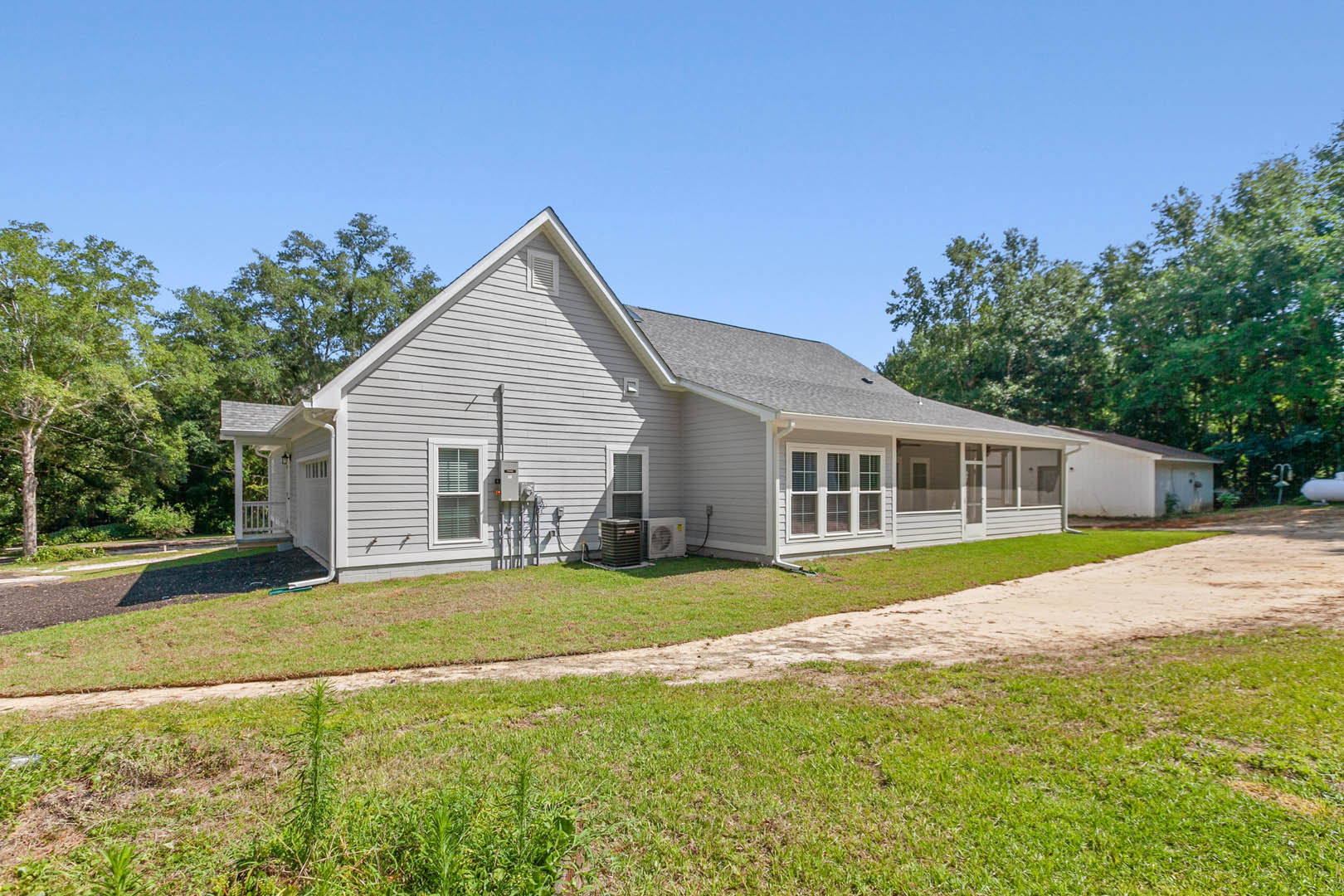 Two-story house with white siding, large windows with blinds, black metal HVAC unit beside driveway, green lawn, and mature trees under blue sky