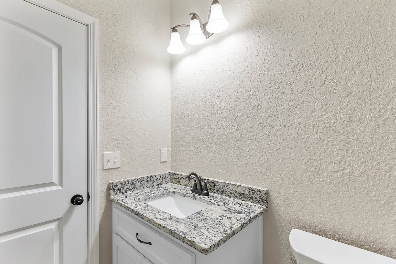 Bathroom with marble countertop, undermount sink with black faucet, white toilet, three-light fixture above mirror, white cabinetry, and light switch on pale wall.
