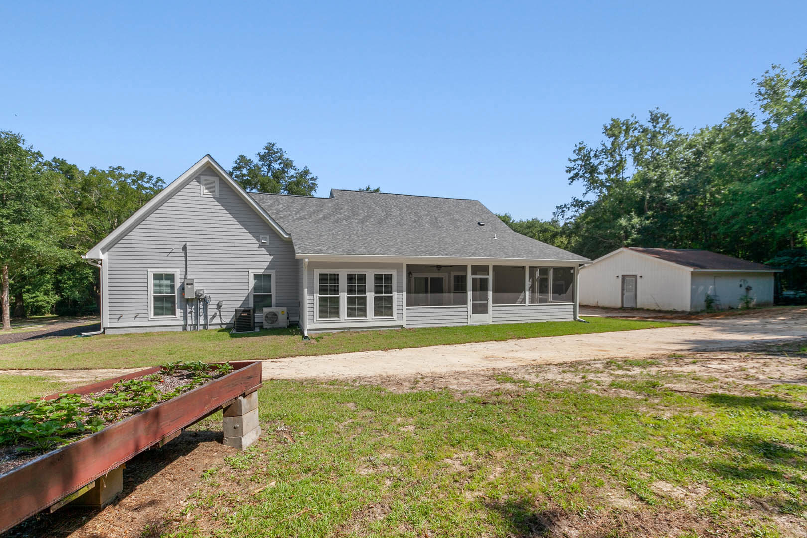Two-story house with covered porch, planter box filled with greenery, dirt path leading to entrance, window with closed blinds, exterior air conditioner unit, white shed with brown