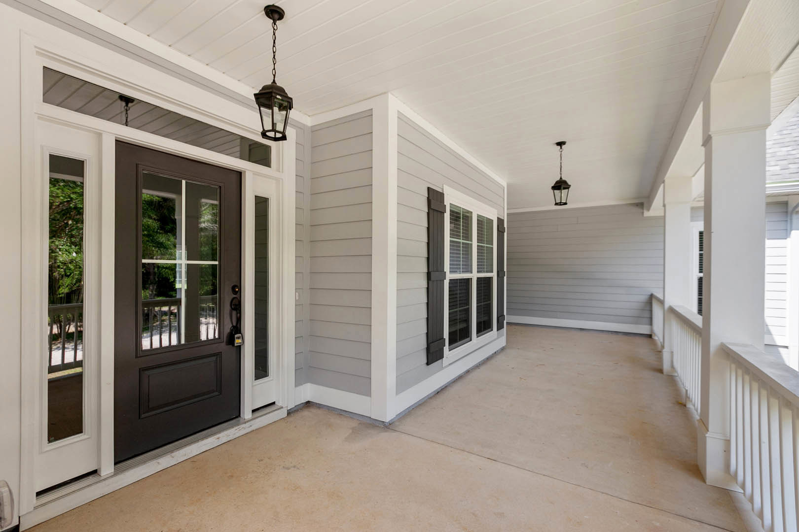 Wide concrete front porch with gray siding, white trim, dark wood door, black metal chain, and modern wall-mounted lamp.