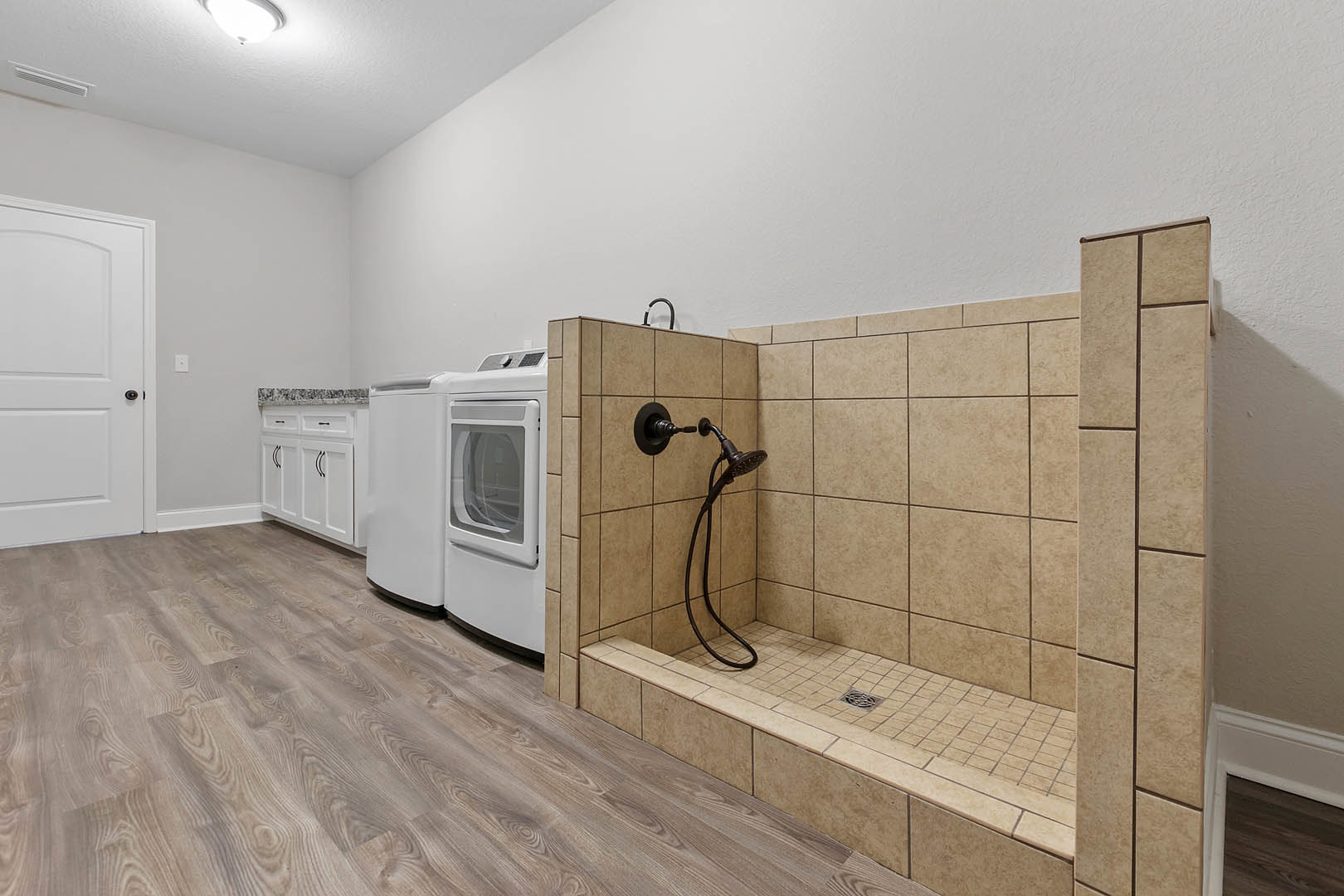 Laundry room featuring a shower with a wall-mounted hose, stacked washer and dryer, white cabinetry, black door handle, white refrigerator beside a window, light switch and fixture