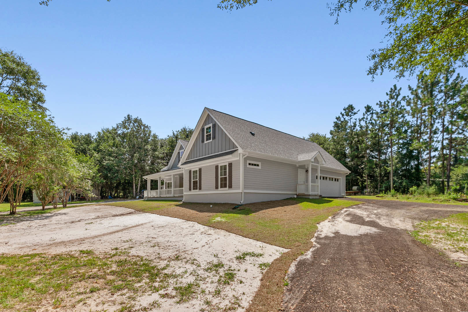 White two-story house with gabled roof, paved driveway, manicured lawn, and mature trees in the background